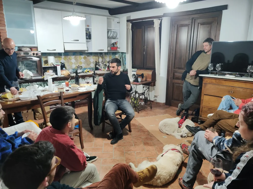 Incontro informale in casa tra i partecipanti al progetto Gruppo di persone riunite in una cucina-salotto tradizionale sarda per un incontro informale serale, sedute in cerchio tra sedie e pavimento con calici di vino rosso, un cane bianco sdraiato al centro, tavolo apparecchiato con cibo sullo sfondo.