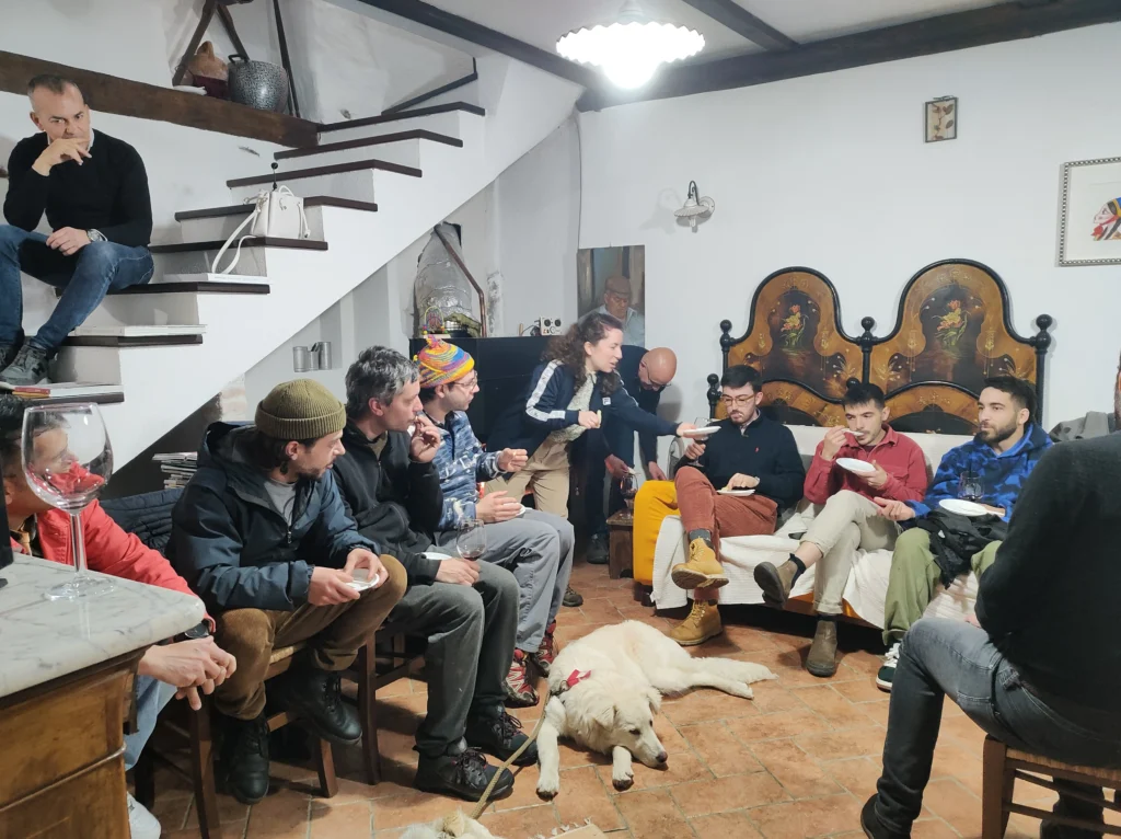 Serata conviviale in una casa tradizionale sarda Gruppo di giovani seduti in un salotto tradizionale sardo con pavimento in cotto, scala in pietra e testiera del letto dipinta, mentre mangiano e conversano in un'atmosfera informale con un cane bianco sdraiato al centro della stanza.