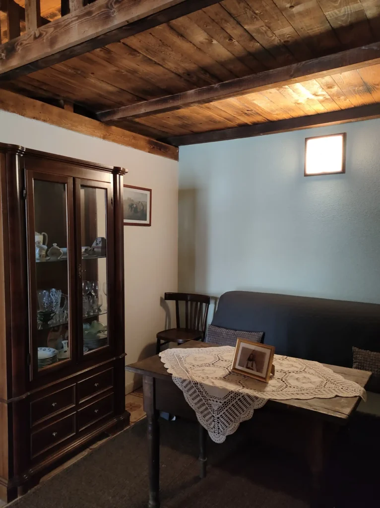 Interno di "Tia Soboa" casa tradizionale sarda con soffitto in legno Stanza di una casa tradizionale sarda con soffitto a travi di legno scuro, credenza in noce con stoviglie in vetrina, tavolo con centrino all'uncinetto e fotografia incorniciata, divano grigio e piccola finestra quadrata sulla parete azzurra.