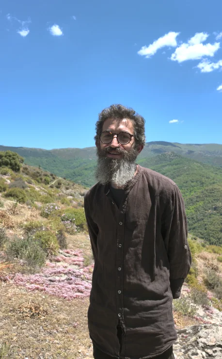 Ledian Agolli a Melanu, Desulo Ledian Agolli, con barba folta, occhiali e camicia scura, in posa sorridente sulla collina di Melanu a Desulo, con fiori di erica rosa, macchia mediterranea e le montagne della Barbagia sotto un cielo azzurro estivo sullo sfondo.