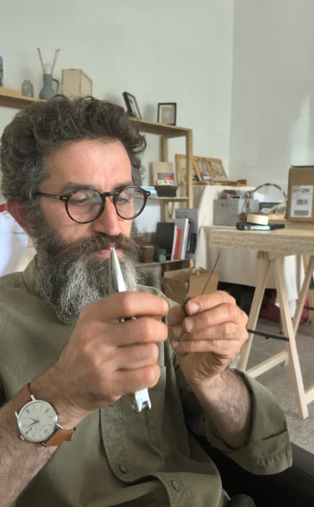 Ledian Agolli al lavoro con il filo di ferro nel laboratorio di Ommo Design Ledian Agolli, con occhiali, barba folta e camicia verde, concentrato mentre piega il filo di ferro con le mani nel laboratorio di Ommo Design a Desulo, con scaffali e materiali creativi sfocati sullo sfondo.