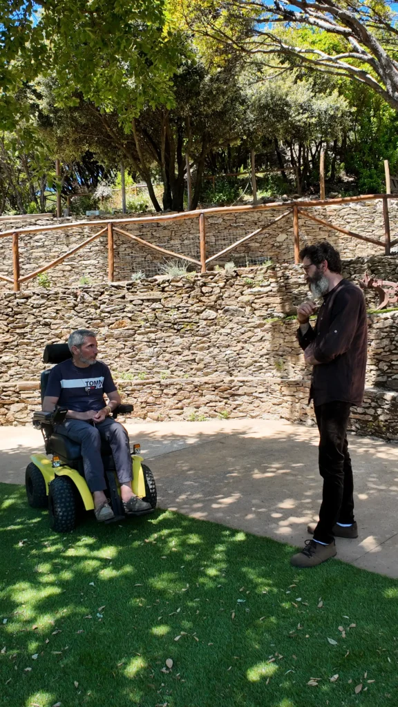Ledian Agolli in visita al parco inclusivo montano Su Dottoreddu de Melanu Ledian Agolli conversa con un uomo su una sedia a rotelle elettrica fuoristrada gialla nel parco inclusivo montano Su Dottoreddu de Melanu, circondati da prato verde, un muretto a secco e alberi sotto un cielo soleggiato.
