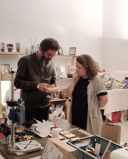 Ommo design e Ledian agolli lavorano assieme nello studio Francesca di Ommo Design Studio e Ledian Agolli esaminano insieme un oggetto in legno tagliato al laser nello studio, circondati da materiali creativi, una stampante 3D e scaffali con oggetti di design sullo sfondo.