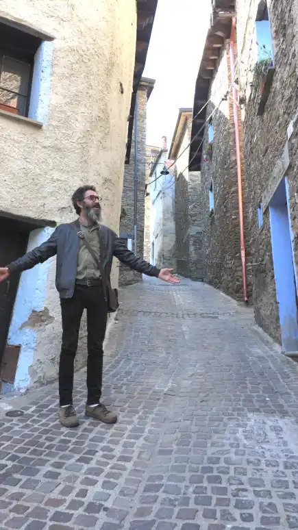 Ledian Agolli in un vicolo di Desulo Ledian Agolli con le braccia aperte in un vicolo lastricato di Desulo, tra case in pietra e intonaco colorato, in un gesto di apertura verso il paese che lo ospita.