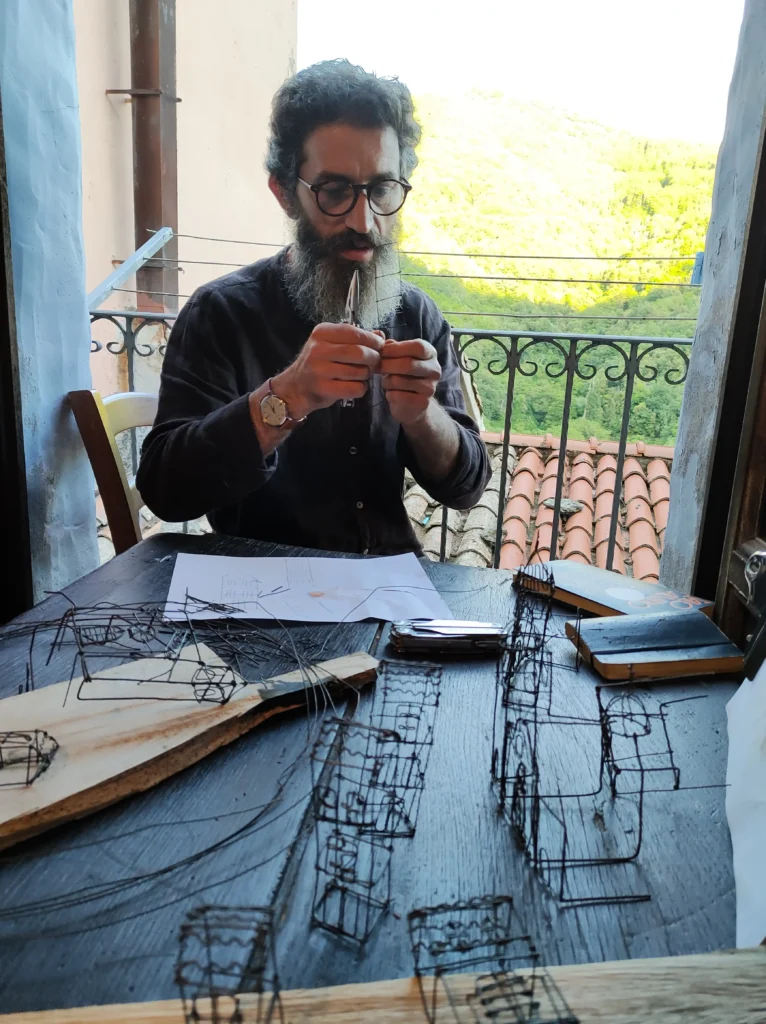 ledian agolli lavora alla sua opera a casetta santa ruge Ledian Agolli, con barba folta e occhiali, concentrato mentre modella il filo di ferro con le mani a un tavolo scuro nella Casetta Santa Ruge, circondato da sculture in progress raffiguranti edifici del paese; sullo sfondo un balcone con tetti in coppi e colline boscose.