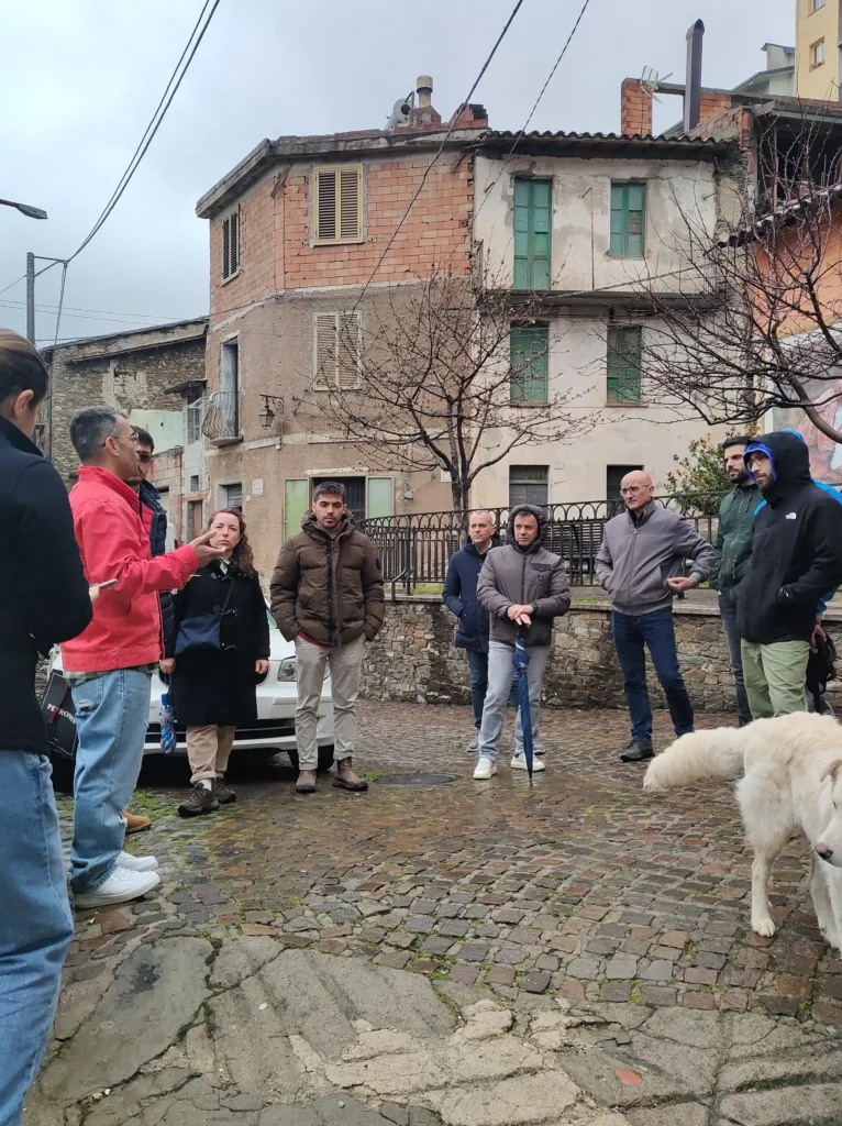 Incontro in piazza sotto la pioggia a Desulo Gruppo di persone in giacche invernali riunite in una piazza lastricata sotto un cielo grigio piovoso, mentre un uomo in giacca rossa parla gesticolando; sullo sfondo edifici storici parzialmente fatiscenti e un cane bianco in primo piano.