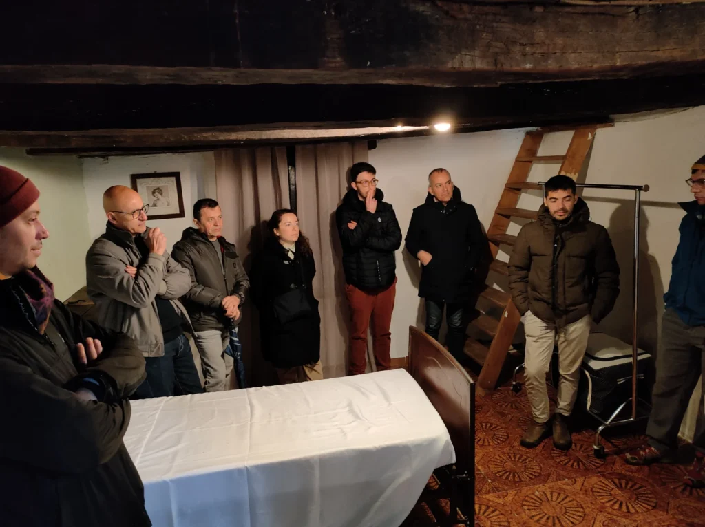 Visita a una casa tradizionale di DesuloVisita a una casa tradizionale di Desulo Visita alla casetta di tia Soboa. Gruppo di persone in piedi in ascolto all'interno di una stanza tradizionale con soffitto a travi scure, pavimento in moquette floreale e scala in legno, durante una visita guidata a una casa storica del paese.